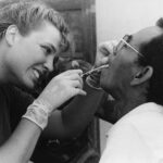 Dental assistant wearing gloves leans in to examine a man's mouth with dental tools during a checkup.