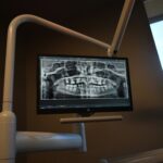 Monitor shows a panoramic dental X-ray of teeth mounted on a white articulating arm in a clinical room