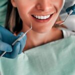 Person in dental chair smiling as dentist in blue gloves uses mirror and scaler near teeth.
