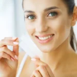 Smiling woman applying skincare with a cotton pad to her cheek near a bright, clean background