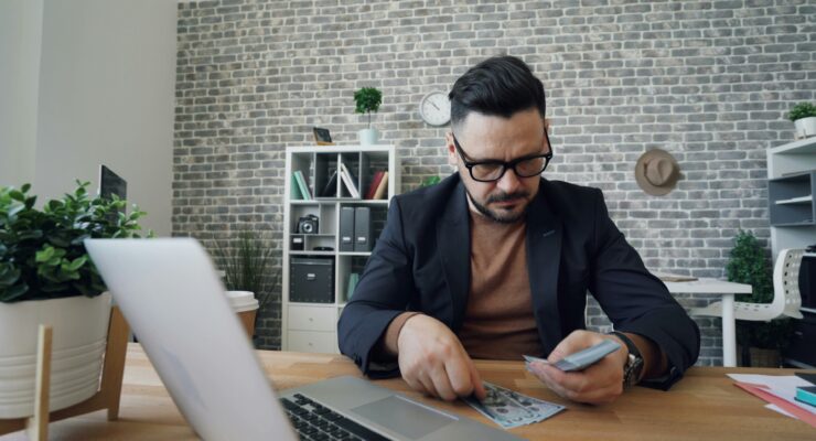 Man in a dark blazer counting cash at a wooden desk in a modern office, laptop open nearby.